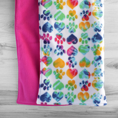 Dog blanket hanging up with cute paw prints and hearts filled with rainbow colors on white background. Back is a solid hot pink color.