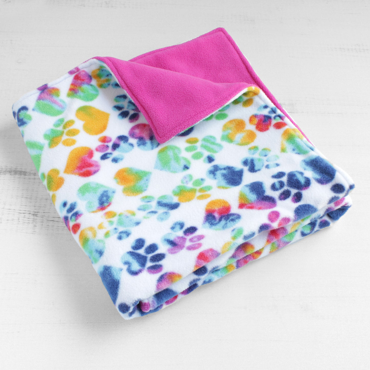 Dog blanket folded up with cute paw prints and hearts filled with rainbow colors on white background. Back is a solid hot pink color.